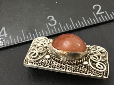 Sale Photo Thumbnail #163: Rectangular silver-tone filigree metal brooch featuring elaborate scrollwork and a central oval carnelian or agate cabochon stone. Measures approximately 1.5 inches in length. Features a classic pin back closure. Shows mild patina and vintage wear consist