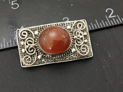 Sale Photo Thumbnail #161: Rectangular silver-tone filigree metal brooch featuring elaborate scrollwork and a central oval carnelian or agate cabochon stone. Measures approximately 1.5 inches in length. Features a classic pin back closure. Shows mild patina and vintage wear consist