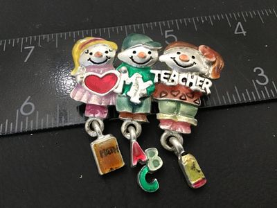 Sale Photo Thumbnail #114: Silver-tone metal brooch featuring three children spelling out "I [Heart] My Teacher" with hanging charms depicting a math book, ABCs, and a pencil. Hand-painted enamel details in various colors. Marked "© AJMC" on the back. Standard pin stem with lockin