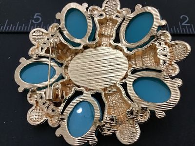 Sale Photo Thumbnail #16: Ornate gold-tone statement brooch pin featuring a large central faux pearl surrounded by teal cabochons, faux pearls, and royal blue rhinestones in a starburst/cross design. Measures approximately 2.5 inches in diameter. Features a pin back closure in wor