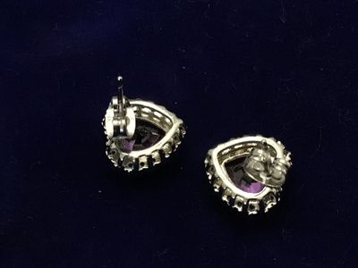 Sale Photo Thumbnail #106: Pair of silver-tone stud earrings featuring heart-cut purple central stones surrounded by a halo of clear accent stones. Secured with friction post backs. Each earring measures approximately 1/2 inch in size. Good condition with light wear.