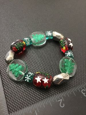 Sale Photo Thumbnail #229: Festive stretch beaded bracelet featuring clear glass beads with green glitter Christmas trees, red beads decorated with white stars and green foliage, green snowflake accent beads, and faceted silver-tone spacer beads. Measures approximately 2.5 to 3 inc