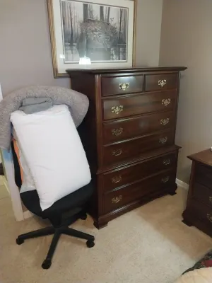 Sale Photo Thumbnail #74: Traditional dark wood tallboy chest of drawers with batwing brass hardware, displayed beside an office chair holding bedding and under a framed wildlife print.