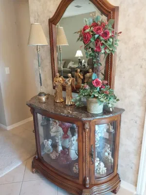 Sale Photo Thumbnail #47: Traditional wooden curio console cabinet with a marble top, accompanied by a matching wall mirror, thin buffet lamp, decorative angel figurines, and floral arrangements.