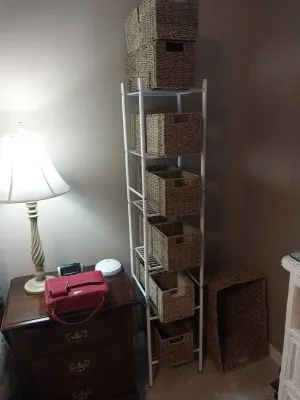 Sale Photo Thumbnail #80: Tall white wire shelving unit filled with woven seagrass storage baskets, shown alongside a dark wood nightstand with a lamp and red bag.