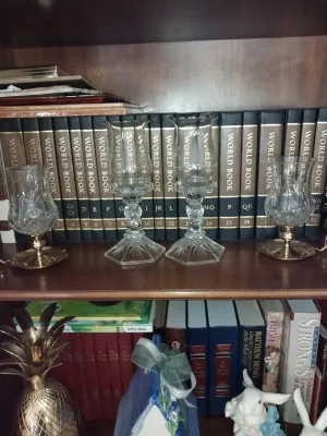 Sale Photo Thumbnail #17: Set of clear cut-glass hurricane lamps and candle holders arranged on a wooden bookshelf in front of a vintage World Book Encyclopedia set.