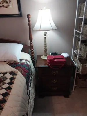 Sale Photo Thumbnail #81: Traditional wooden three-drawer nightstand alongside a four-poster bed frame, topped with a cream-colored spiral table lamp and red organizer bag.