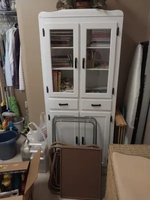 Sale Photo Thumbnail #97: Tall white painted storage cabinet with upper glass doors, central drawers, and lower cabinet doors, shown alongside folded metal snack tables.