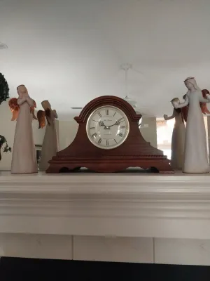 Sale Photo Thumbnail #11: Traditional wooden mantel clock with Roman numerals, flanked by decorative angel figurines on a mantelpiece.