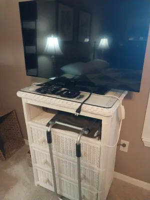 Sale Photo Thumbnail #77: White painted wicker dresser with glass top, open shelf, and three drawers, topped with a flat-screen television and remotes.