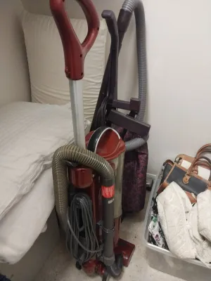 Sale Photo Thumbnail #88: Red upright bagless vacuum cleaner stored next to a bed and a plastic tub filled with handbags and textiles.
