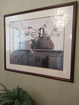 Sale Photo Thumbnail #48: Framed still life art print depicting flowering branches in a woven basket atop a wooden cabinet, housed in a mahogany-toned frame.