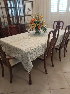 Sale Photo Thumbnail #33: Traditional Queen Anne style wooden dining table set with matching high-back chairs, covered with a white lace tablecloth and centered with a floral arrangement.