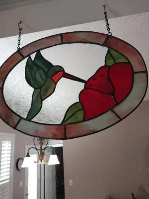 Sale Photo Thumbnail #20: Oval stained glass suncatcher featuring a hummingbird sipping nectar from a red flower, suspended from black hanging chains.