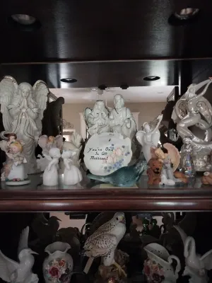 Sale Photo Thumbnail #13: Collection of white porcelain and ceramic angel figurines, decorative plaques, and bird sculptures arranged inside a dark wooden display cabinet.