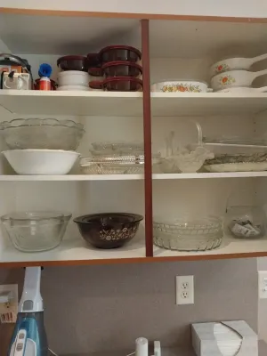 Sale Photo Thumbnail #25: Collection of vintage glassware and cookware, including CorningWare, Pyrex, and clear pressed glass serving pieces arranged on cabinet shelves.