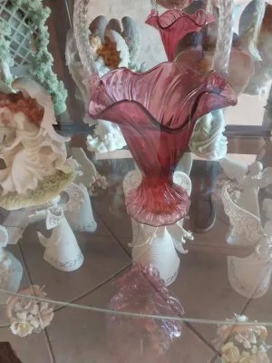Sale Photo Thumbnail #45: Vintage cranberry glass basket with a ruffled rim and clear applied handle, displayed in a glass cabinet alongside various white angel figurines.
