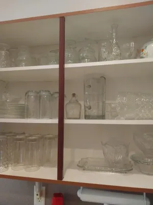 Sale Photo Thumbnail #31: Collection of vintage clear glassware displayed on white shelving, including drinking glasses, cut crystal pieces, pitchers, and serving trays.
