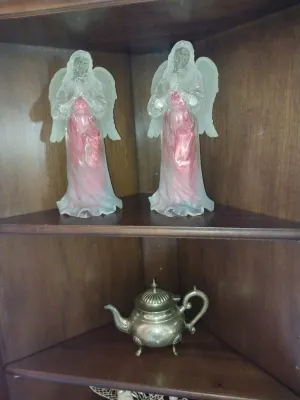 Sale Photo Thumbnail #52: Pair of frosted glass angel figurines with pink accents displayed above a small vintage silver-plated teapot on a wooden corner shelf unit.