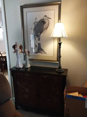 Sale Photo Thumbnail #10: Large framed blue heron print hanging above a dark wooden console table with metallic-winged angel figurines and a slim table lamp.