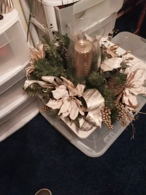 Sale Photo Thumbnail #61: Festive holiday floral centerpiece featuring artificial greenery, metallic gold ribbon, poinsettias, and a clear glass hurricane vase holding a gold pillar candle.