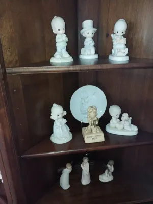 Sale Photo Thumbnail #16: Collection of Precious Moments porcelain figurines, Willow Tree angel figures, and a decorative plate displayed inside a dark wood corner cabinet.