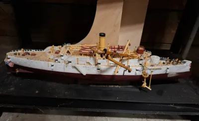 Detailed scale model of a pre-dreadnought battleship or protected cruiser on a display stand, featuring fine rigging, lifeboats, and deck...