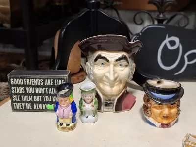 Collection of vintage character jugs and ceramic figurines displayed alongside a wooden sign with a quote.