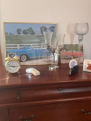 Sale Photo Thumbnail #17: Collection of tabletop decor including framed '57 Chevy Bel Air prints, a brass alarm clock, a three-arm glass candelabra, and a tall blown glass goblet arranged on a wooden dresser.