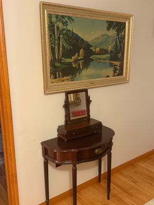 Sale Photo Thumbnail #18: Traditional mahogany demi-lune console table with brass hardware, set below a framed mountain landscape print and topped with a small vanity mirror with a single drawer.