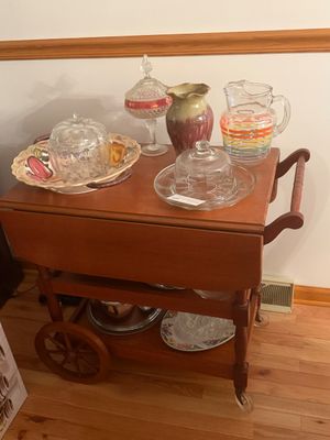 Sale Photo Thumbnail #14: Vintage drop-leaf wooden tea cart on spoked wheels, featuring a top display of various glassware, a colorful striped pitcher, ceramic vase, and serveware.
