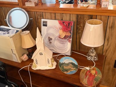 Sale Photo Thumbnail #22: Collection of tabletop decor including an illuminated porcelain church sculpture, table lamps, a vanity mirror, a Mikasa server in box, and religious wall plaques.