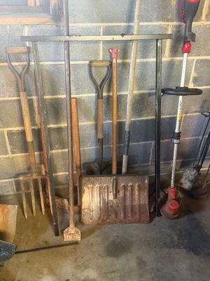 Sale Photo Thumbnail #8: Collection of assorted garden and yard tools including shovels, pitchfork, ice scraper, and Milwaukee weed trimmer leaning against a cinderblock wall.