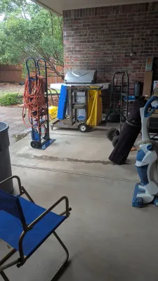 Covered patio containing hand trucks, extension cords, a stainless steel gas grill, a janitorial cart, folding chairs, and a vacuum cleaner.