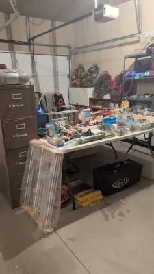 Garage sale setup featuring a brown metal four-drawer filing cabinet, folding tables filled with miscellaneous household items, and...