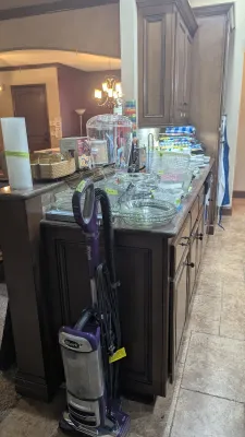 Purple Shark upright vacuum standing beside a kitchen counter filled with glassware, kitchenware, and household items.