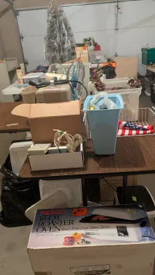 A collection of household goods and appliances arranged on folding tables in a garage, featuring a boxed Nesco 18-quart roaster oven in...