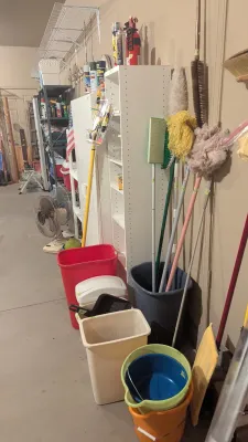 Assorted household cleaning supplies, including mops, brooms, buckets, and plastic trash cans alongside shelving units.