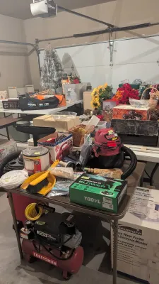 Garage sale setup featuring a red Craftsman air compressor, a Shop-Vac HangUp Mini, a boxed Yoovl handheld blower, artificial flowers,...