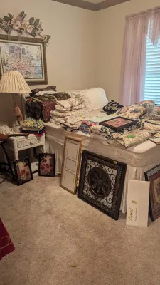 Sale Photo Thumbnail #13: Bedroom featuring a bed stacked with assorted quilts and linens, flanked by white wicker furniture, table lamps, and framed wall art leaning against the base.