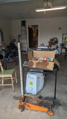Sale Photo Thumbnail #29: Vintage heavy-duty orange hydraulic floor jack displayed on a garage floor, alongside folding tables filled with miscellaneous household items and tools.