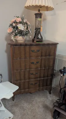 Sale Photo Thumbnail #14: French Provincial style four-drawer wooden dresser with ornate metal bail handles, topped with a decorative table lamp and artificial floral arrangement.