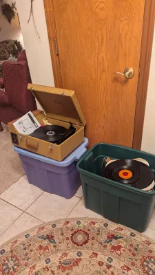 Sale Photo Thumbnail #9: Vintage-style Crosley tweed suitcase record player resting on a plastic bin alongside a green storage container filled with vinyl records.