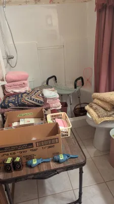 Sale Photo Thumbnail #15: Assorted bathroom items and linens displayed on tables, including folded towels, bath mats, decorative bird figurines, and a medical shower chair inside the stall.