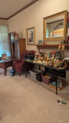 Sale Photo Thumbnail #6: Living room setup featuring a folding table covered in decor items, a burgundy wingback armchair, an ornate gilt framed mirror, and wooden furniture.