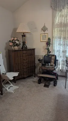 Sale Photo Thumbnail #22: Four-drawer dark wood dresser in French Provincial style, shown alongside a table lamp, artificial floral arrangements, and a motorized mobility chair.