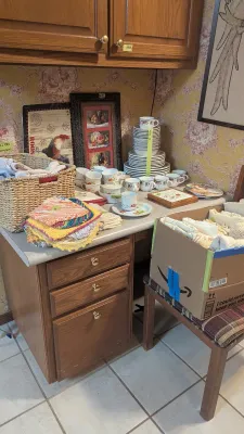 Sale Photo Thumbnail #36: Kitchen counter display featuring a stacked dinnerware set with floral motifs, crocheted potholders, framed wall art, and a cardboard box of linens.