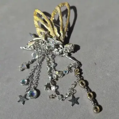 Sale Photo Thumbnail #112: Whimsical fairy or angel brooch featuring textured gold-tone wings, rhinestone-embellished star accents, and dangling bezel-set crystal chains, dated 2000.