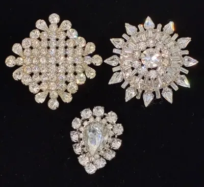 Sale Photo Thumbnail #91: Trio of vintage rhinestone brooches in silver-tone settings, including diamond, starburst, and teardrop designs.