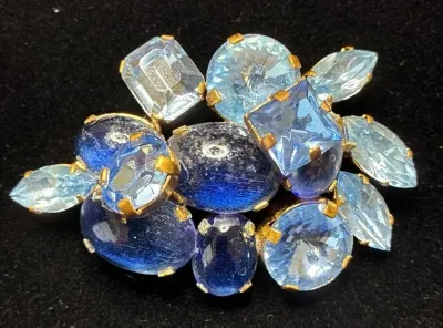 Sale Photo Thumbnail #81: Vintage gold-tone brooch featuring an arrangement of light and dark blue rhinestones and cabochons in various cuts, including emerald, marquise, and oval shapes.